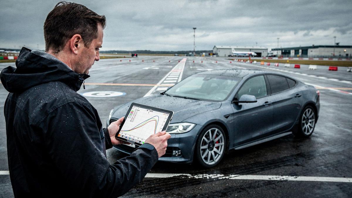 Instructor beside a sedan in a wet airfield braking lane, holding a tablet with a telemetry trace on screen